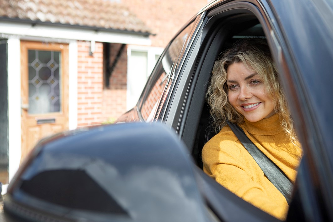 A woman with blonde curly hair, wearing a yellow turtleneck sweater and a seatbelt, smiles from the drivers seat of a black car. The side mirror of the car is visible in the foreground, and a brick house with a wooden door is in the background.