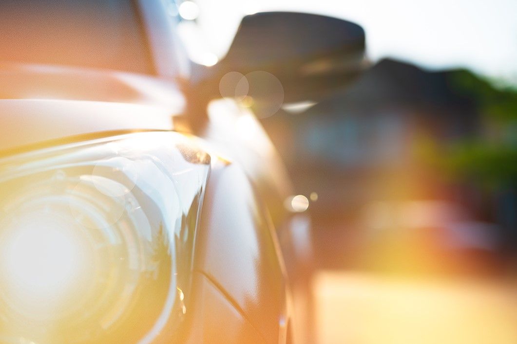 Close-up of a car headlight with sunlight creating lens flare effects. The background is blurred, emphasizing the bright and shiny surface of the vehicle.