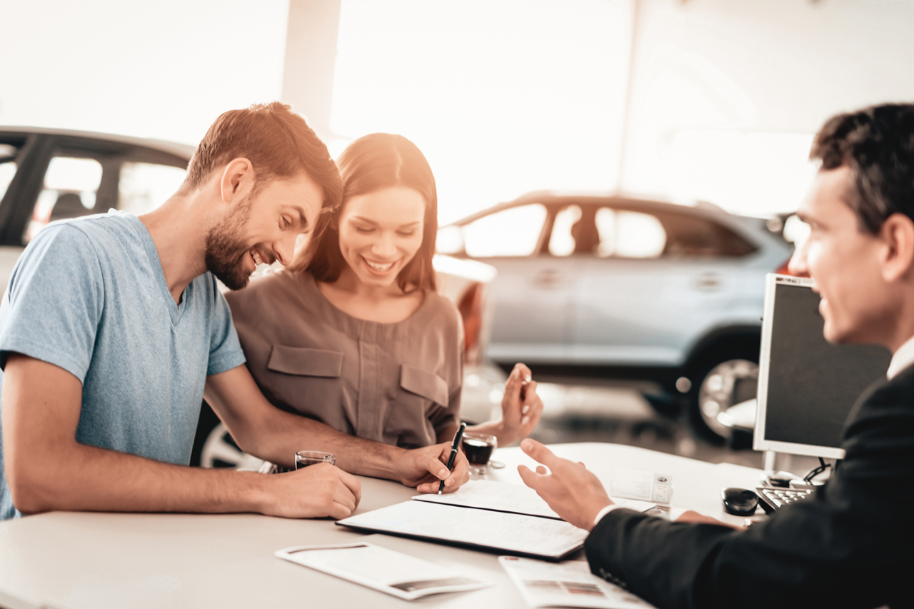 Happy couple signing paperwork at a car dealership with a sales agent.