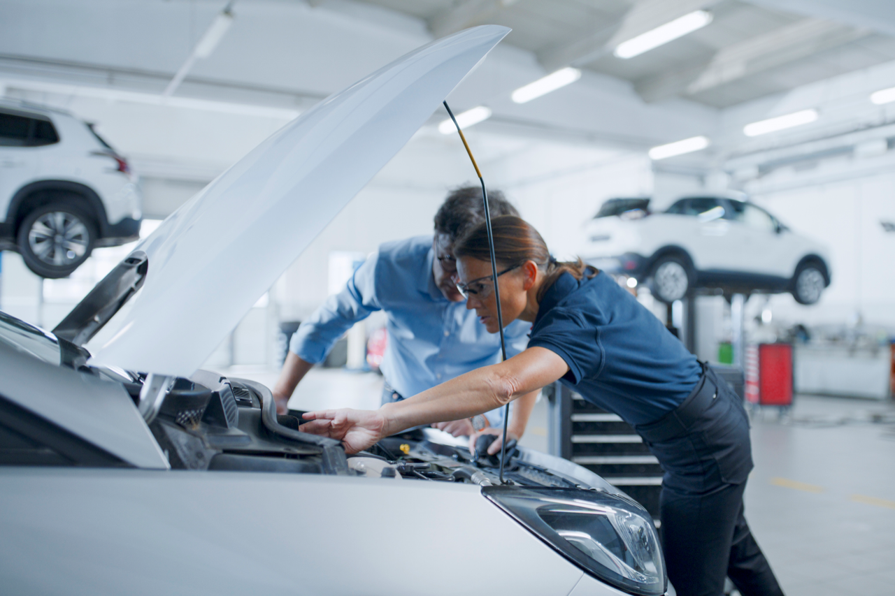 Two mechanics inspecting the engine of a car with the hood open in an auto repair shop. Another car is lifted on a hydraulic lift in the background.